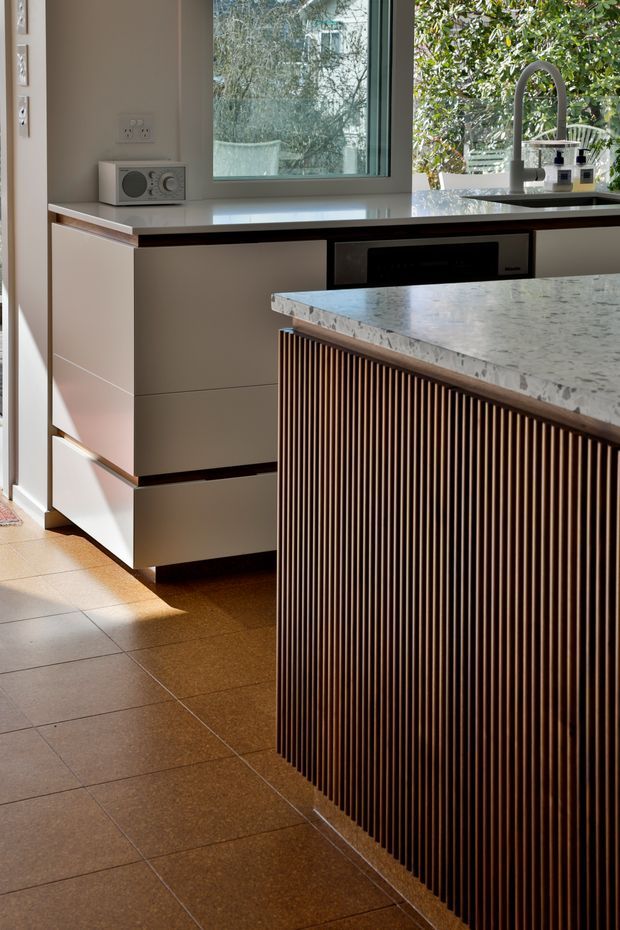 A contemporary kitchen focused on a central island with a pale stone benchtop and a vertically fluted dark timber front. Behind it, streamlined light-coloured cabinetry incorporates an oven, while a dark gooseneck tap and sink sit beneath a large window overlooking greenery. Warm tiled flooring and natural daylight create a refined, inviting atmosphere.