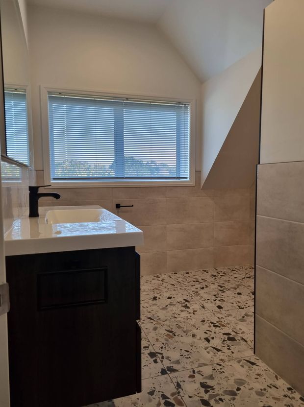 A contemporary bathroom with a dark timber vanity, white integrated basin and matte black tapware. The room features large beige wall tiles, a terrazzo-style floor, a frameless glass shower screen, and a broad window with horizontal blinds. The sloped ceiling and angular shower enclosure add architectural interest.