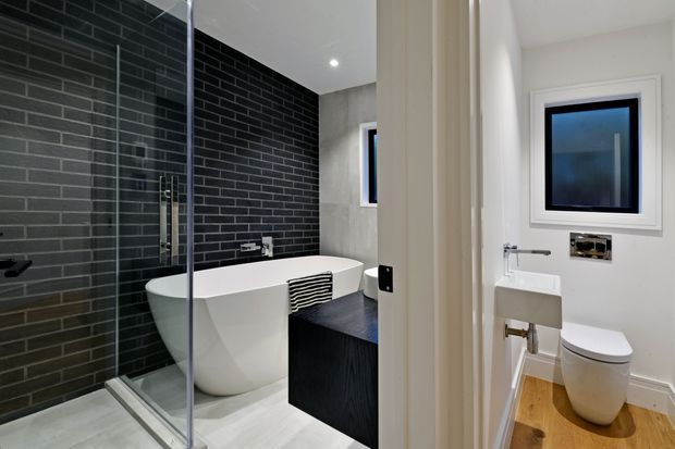 A contemporary bathroom view showing a walk-in glass shower, freestanding white bathtub, dark charcoal brick-look tiled feature wall, black vanity, and a separate toilet area visible through the opening. The space combines white walls and fixtures with dark tile accents and warm timber flooring in the toilet zone.