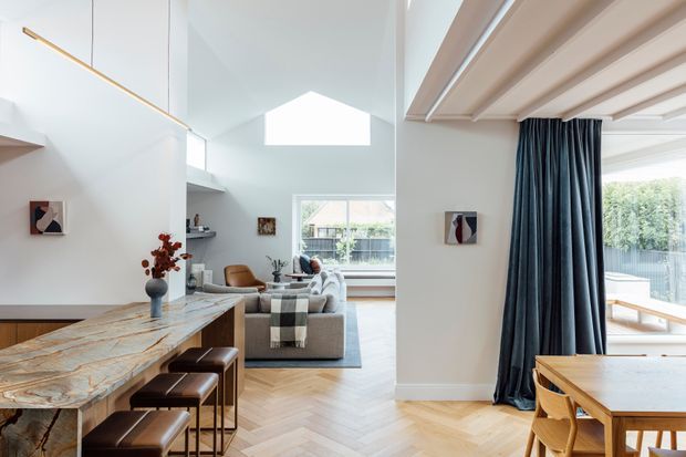 Bright, open-plan contemporary interior combining a kitchen and dining area with a living room. The space features a veined stone island with dark stools, pale herringbone timber flooring, white walls, a high ceiling with clerestory-style glazing, exposed timber ceiling beams, and full-height dark curtains separating the living and dining zones. Large windows bring in abundant natural light and show greenery outside.