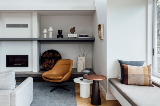 A bright, refined living room with a built-in fireplace wall, dark stone or concrete hearth and shelving, decorative objects, a tan leather lounge chair, small side tables, and a window seat with layered cushions. Light walls, pale timber flooring, and soft daylight create a calm contemporary setting.