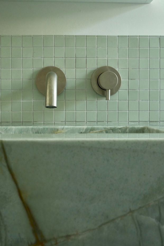 Close-up of a minimalist bathroom wall featuring brushed metal wall-mounted tapware and a separate circular control set into a pale green square-tiled splashback. A thick stone or marble ledge and tub surround occupy the lower portion, with visible veining and some aged patina.