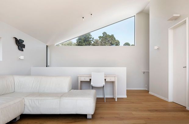 Bright minimalist living room with a large white sectional sofa, compact white writing desk and chair, pale walls, warm timber flooring, and a high triangular clerestory window overlooking trees. A hallway and doorway are visible on the right.