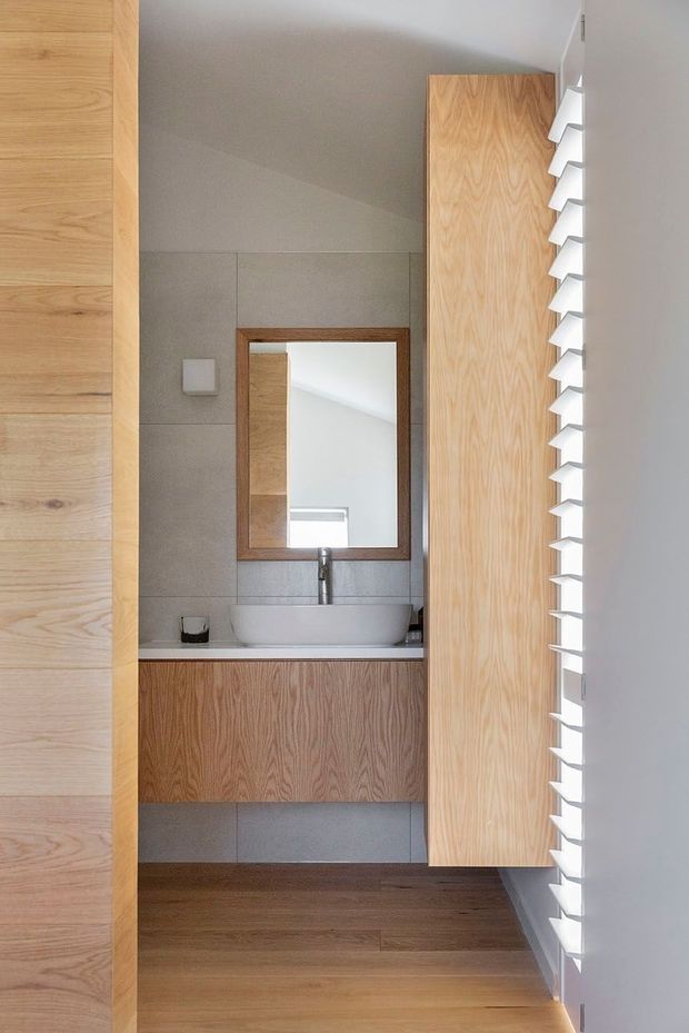 Compact contemporary bathroom viewed through a timber-lined doorway, featuring a floating timber vanity with a white vessel basin, chrome tapware, a timber-framed mirror, large pale grey wall tiles, timber flooring, and a tall bank of white horizontal louvres providing filtered daylight.