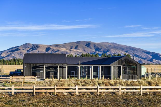 A modern single-storey rural house set behind timber post-and-rail fencing, with a dark metal gabled roof, dark vertical cladding, extensive glazing, and a covered carport or entry structure. Dry grassland and agricultural fields surround the home, with a prominent mountain range in the background.