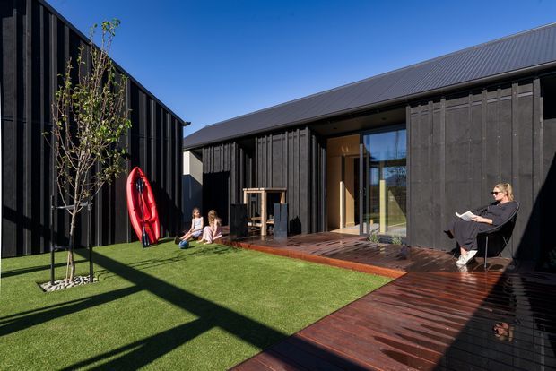 A contemporary dark-clad residential courtyard with a strong L-shaped composition. Vertical black timber or composite cladding encloses the courtyard, with a low pitched metal roof, large glazed doors, and a dark timber deck surrounding a lawn area. A red surfboard, children, and a seated adult add signs of active family use. Strong sunlight creates pronounced structural shadows across the lawn and deck.
