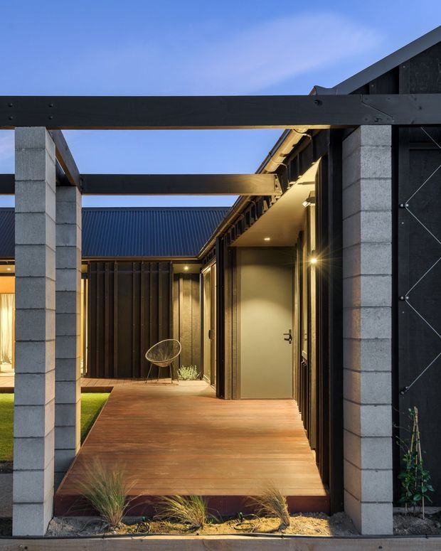 An evening exterior view of a contemporary courtyard entry framed by dark steel beams and pale concrete block columns. A timber deck walkway leads past a recessed, warmly lit entry door and dark metal-clad walls, with exposed overhead beams, a pitched roofline, and small areas of ornamental planting visible around the perimeter.