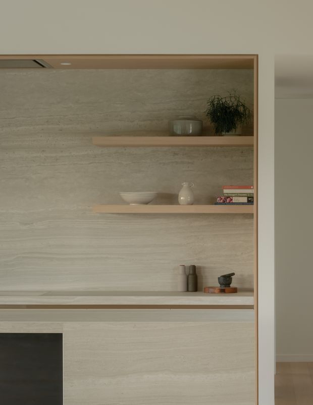 A close-up of a refined built-in shelving and cabinetry feature. The unit has a full-height pale beige stone-look textured wall surface, integrated timber-toned shelves, slim matching trim, and lower cabinetry. Minimal ceramics, books, and dark decorative vessels are arranged on the shelves. A partial opening at right reveals an adjacent neutral interior with timber flooring. The overall composition is quiet, warm, and minimalist.
