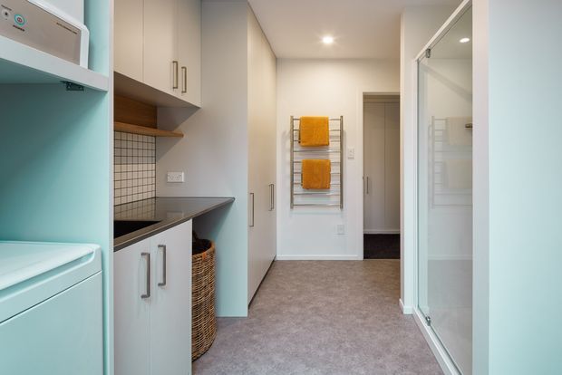 Compact, bright laundry room with pale cabinetry, a dark benchtop, tiled splashback, stacked appliances, open shelving, and a wall-mounted heated towel rail with orange towels. A full-height glass door on the right opens to a shower or wet-area enclosure, while a doorway leads to an adjacent hall or room.
