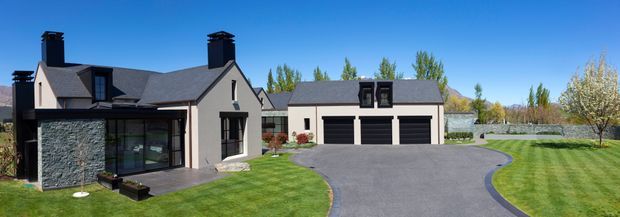 Exterior view of a large contemporary gabled home with dark grey rendered walls, steep pitched roofs, black-framed glazing, and a detached multi-bay garage. The property is set within a broad landscaped lawn with a curved paved driveway and mature trees under a clear blue sky.