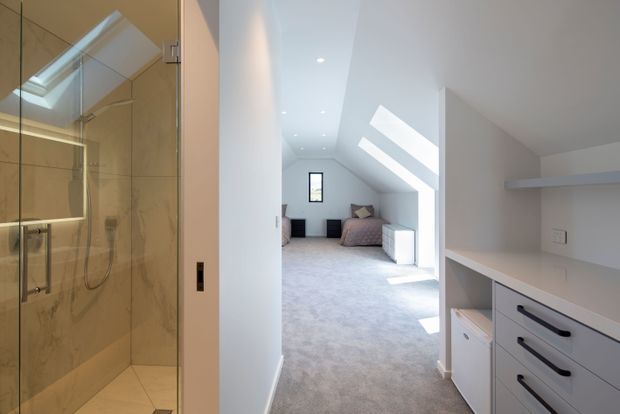 Bright contemporary upper-level interior showing a carpeted bedroom beneath a sloped ceiling with skylights and strong natural light. A compact white built-in study or storage joinery area occupies the right foreground, while a glass-enclosed tiled shower is visible to the left.
