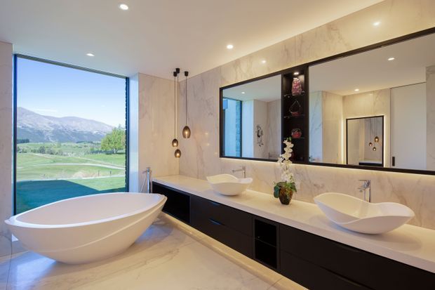 A spacious contemporary bathroom with a freestanding white bathtub positioned beside full-height glazing overlooking a green landscape and mountains. A long double vanity with white vessel basins, dark built-in cabinetry, a pale stone countertop, large marble-look wall panels, and a wide mirrored cabinet dominate the room. Recessed ceiling lights and pendant lights provide warm, refined illumination.