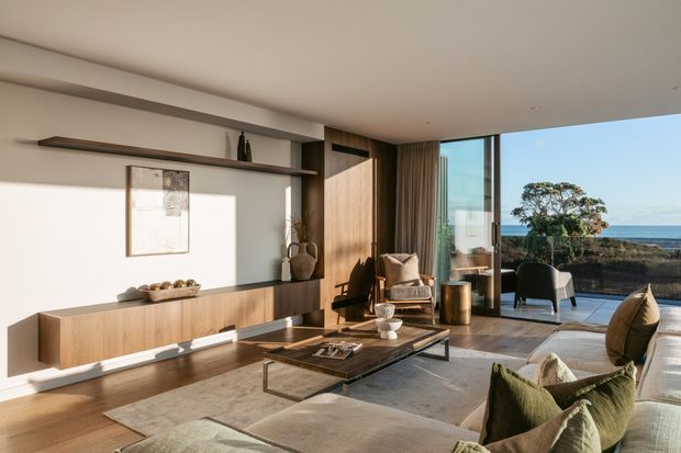 A refined contemporary living room with warm timber built-in cabinetry and open display shelving, a neutral plastered wall, large area rug, low rectangular coffee table, and upholstered lounge seating. Full-height glazing opens to a terrace with an ocean view, bringing in strong natural light. The room has a warm, calm, coastal outlook with restrained modern styling.