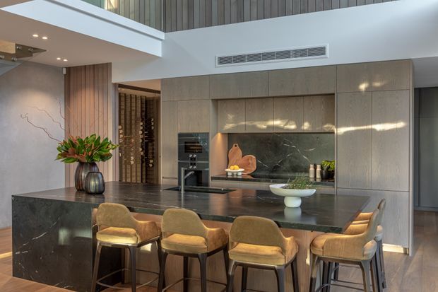 A sophisticated open-plan kitchen with a large dark stone island and integrated seating for several people. Full-height timber-toned cabinetry lines the rear wall, framing a dark veined stone splashback and built-in appliances. Warm upholstered bar stools, subtle under-cabinet lighting, and a double-height ceiling with a vertical timber feature wall create a refined, contemporary interior.