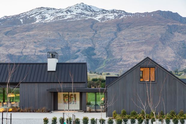 Exterior view of a contemporary dark-clad residential property composed of steep gabled and pitched-roof forms, with large glazed openings and warm timber-framed windows. The home sits against a dramatic alpine mountain backdrop, with landscaped planting and a paved foreground.