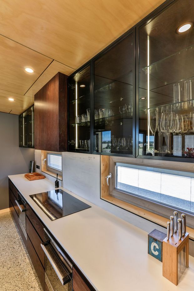 A compact, refined kitchen with a long pale stone benchtop, dark timber cabinetry, black-framed glass-fronted overhead cupboards displaying glassware, integrated appliances, and a black induction cooktop. Warm timber ceiling panels and window surrounds contrast with the cool grey wall and terrazzo-style floor. Under-cabinet and recessed lighting create a polished contemporary atmosphere.