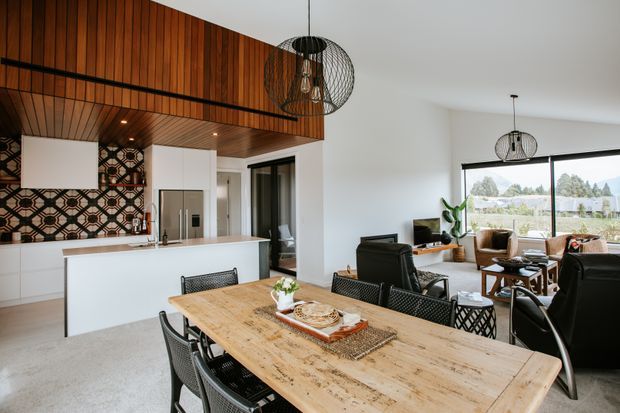Open-plan kitchen, dining, and living area with a white kitchen, stainless-steel appliances, patterned tile splashback, timber cabinetry and ceiling lining, a large timber dining table, black wire-frame pendant lights, and a bright living zone opening to expansive windows.