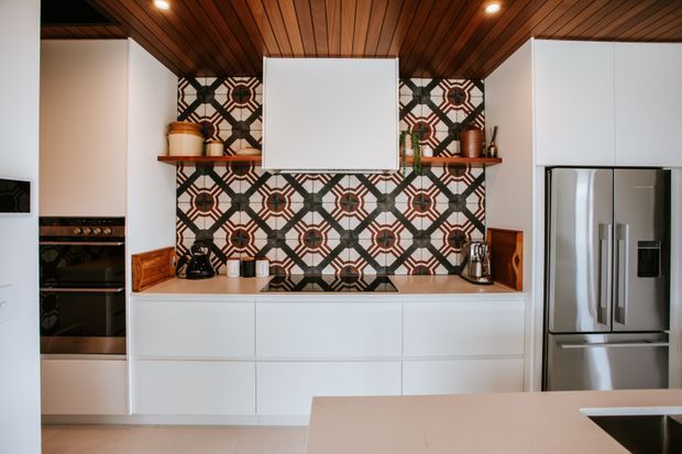 Modern kitchen with white flat-panel cabinetry, integrated appliances, a stainless-steel French-door refrigerator, a patterned geometric tile backsplash, and a warm timber-lined ceiling with recessed lighting. A pale island bench occupies the foreground.