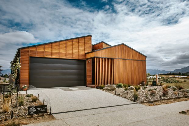 Exterior view of a contemporary timber-clad garage or residential outbuilding with a large dark sectional garage door, pitched and asymmetrical roof forms, and a pale concrete driveway. The building sits in a dry, landscaped setting with gravel, low planting, fencing, and distant mountains visible beneath a dramatic sky.