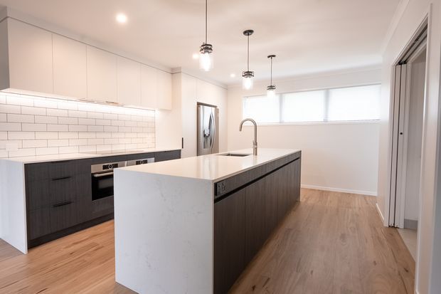 Bright contemporary kitchen with a large waterfall-edge island, dark timber-look lower cabinetry, white upper cabinets, a white subway-tile splashback, integrated appliances, pendant lighting and light timber flooring. A wide horizontal window and open doorway add natural light.