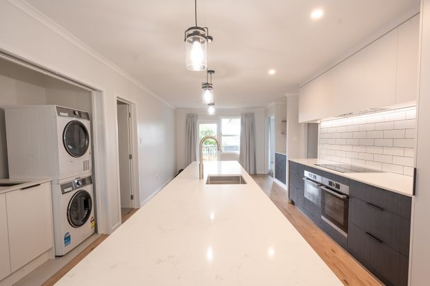 Bright contemporary galley kitchen with a large white stone island, integrated appliances, dark lower cabinetry, white upper cabinetry, and a white subway-tile splashback. A stacked washer and dryer are housed in cabinetry on the left, while pendant lights and recessed lighting illuminate the space. Timber flooring runs through the kitchen toward a glazed opening at the far end.