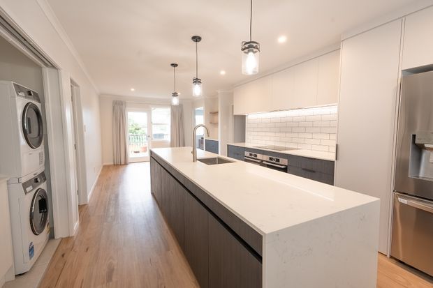 Bright open-plan kitchen viewed from the adjoining hallway, featuring a large island with a pale stone benchtop, dark vertical-grain cabinetry, white cabinetry, white subway-tile splashback, stainless-steel appliances, timber flooring, pendant lights, and a laundry nook with stacked washer and dryer. A dining or living area and glazed opening to the outdoors are visible beyond the kitchen.