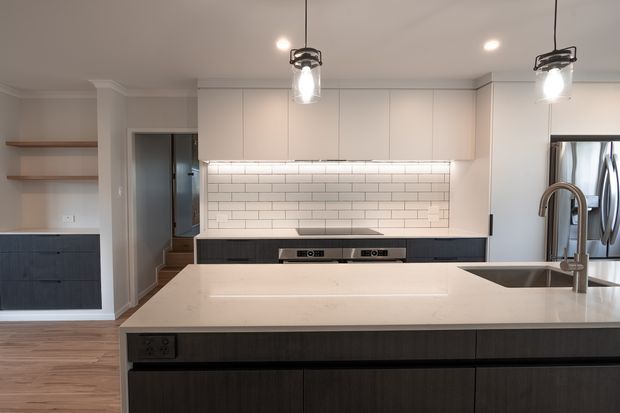 Contemporary kitchen viewed from behind a large island, featuring dark wood cabinetry, a pale engineered-stone benchtop, white subway-tile splashback, integrated stainless-steel appliances, open timber shelves, and pendant lighting. The room has a warm neutral palette and clean, functional detailing.