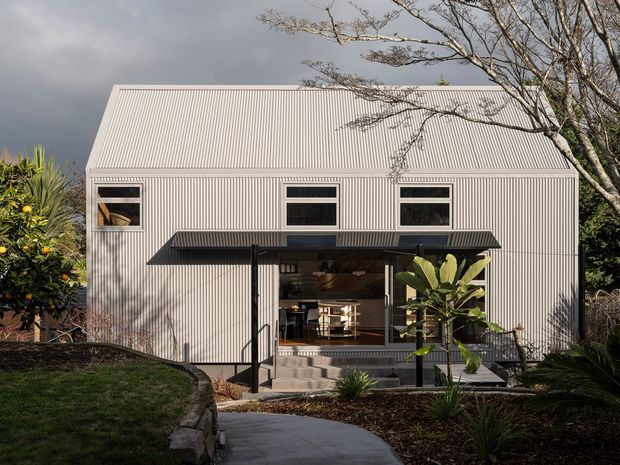 Front exterior view of a compact contemporary two-storey dwelling with a steep gabled corrugated metal roof and vertically ribbed light-coloured metal cladding. The facade has three upper-level windows and a large glazed opening at ground level beneath a dark projecting awning, revealing a warm-toned interior. A concrete pathway, steps, planted garden beds and mature vegetation lead to the entrance.