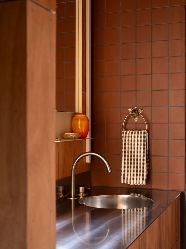 A compact, richly coloured bathroom vanity viewed through a partially open timber door or partition. The space features reddish-brown square tiled walls with dark grout, warm timber cabinetry and shelving, a polished dark benchtop, a brushed metal oval vessel sink, and a curved metal tap. A decorative brass towel ring with a patterned hand towel and a small amber vessel add detail.