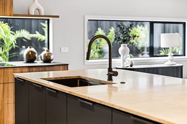 Contemporary kitchen interior with a large island featuring a pale stone benchtop, integrated sink, and tall dark curved tap. Dark cabinetry contrasts with timber cabinetry and a timber-toned counter at the rear. Black-framed windows bring in views of lush planting, while sculptural white ceramics and lamps add restrained decorative detail.