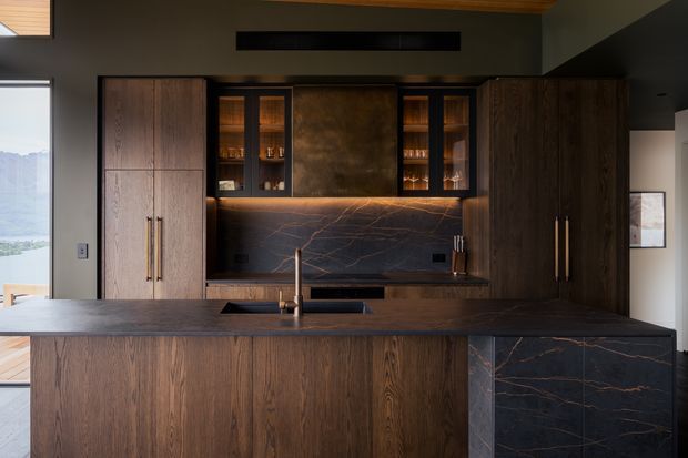 A sophisticated contemporary kitchen with a large dark timber island and full-height timber cabinetry. The island and splashback feature dark stone with dramatic fine veining, while warm integrated lighting illuminates open display shelving and the back benchtop. A large window provides a view to the landscape and water beyond.