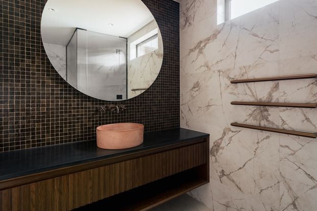 A refined contemporary bathroom vanity featuring a dark mosaic-tile wall, large round mirror, black benchtop, timber cabinetry, and a terracotta vessel basin. The adjacent wall is clad in dramatic veined marble with minimalist timber towel rails, and a fluted-glass shower enclosure is reflected in the mirror.