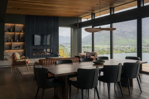 A sophisticated open-plan living and dining area with a long dark timber dining table surrounded by upholstered black dining chairs. The room features a dark vertical timber fireplace wall with integrated shelving and a television, a sculptural pendant light, leather seating, a timber-lined ceiling, and extensive floor-to-ceiling glazing overlooking a dramatic mountain and valley landscape. The palette is dark, warm, and refined, with strong indoor–outdoor connection.