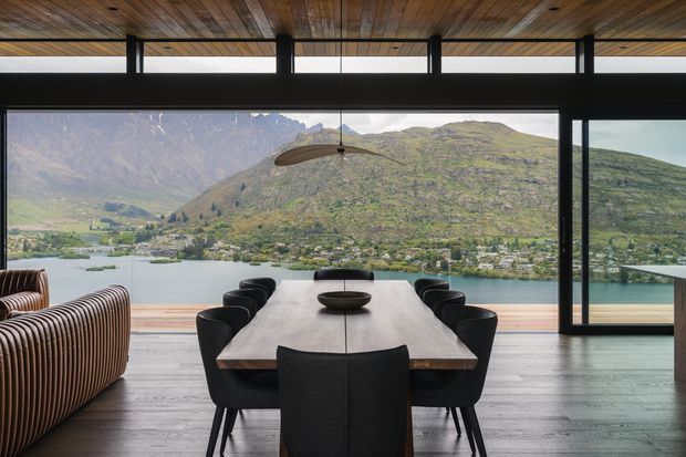 A refined dining area with a long timber dining table and black upholstered chairs, opening through extensive black-framed glazing to a dramatic lakeside and mountainous landscape. The room features a warm timber-lined ceiling, dark timber flooring, a sculptural pendant light, and a large striped upholstered lounge chair in the foreground.