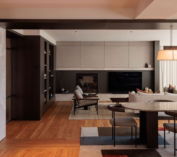 A refined open-plan living and dining area with warm timber flooring, dark built-in cabinetry, a stone fireplace surround, and integrated media wall. The room features a neutral palette, contemporary furnishings, a large round dining table, lounge seating, layered rugs, and pendant lighting. A broad dark ceiling beam and full-height curtains add architectural definition.