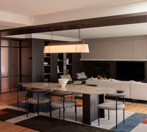 Open-plan contemporary dining and living area with a large oval stone dining table, upholstered dining chairs, a linear pendant light, dark timber joinery and beams, a fireplace wall, and a neutral sectional sofa. Warm timber flooring and layered rugs create a refined, comfortable interior.