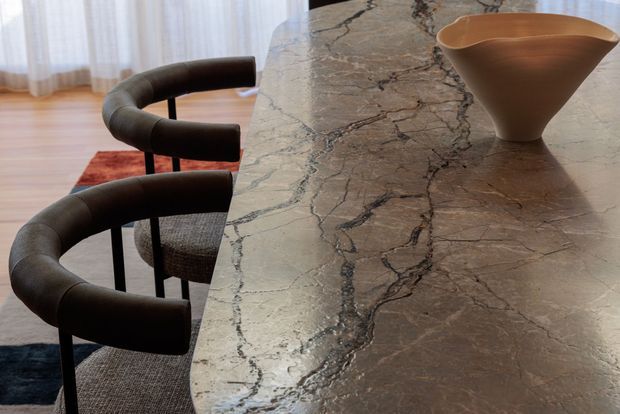 Close-up view of a refined dining area centered on a large stone dining table with dramatic dark veining. Two dark upholstered curved-back dining chairs sit alongside the table, with a sculptural terracotta-toned vessel on top. Sheer curtains, timber flooring, and layered rugs are visible in the background, creating a warm, sophisticated interior.