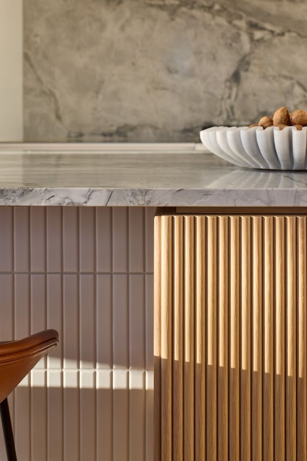Close-up interior detail of a refined built-in joinery unit or kitchen island. The composition features a pale veined stone benchtop, vertically fluted warm timber cabinetry, and muted taupe vertical wall tiles. A sculptural white bowl with nuts sits on the benchtop, while part of a brown leather or timber chair appears at the lower left. Strong sunlight creates crisp shadows across the surfaces.