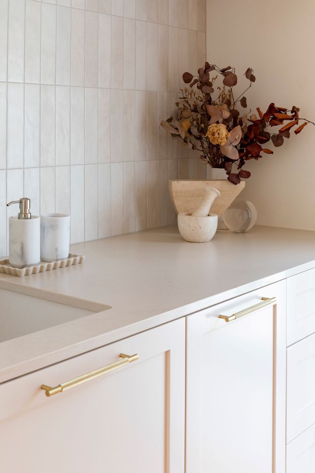 Close-up of a refined light-toned kitchen or bathroom vanity area, featuring white shaker-style cabinetry with brushed brass handles, a pale stone benchtop, and a vertically tiled backsplash. A sculptural stone vessel and dried floral arrangement provide warm decorative accents, while soap dispensers sit beside the partially visible sink.