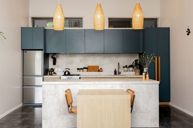 A contemporary galley-style kitchen with dark blue-green full-height cabinetry, a stainless-steel refrigerator, pale stone benchtops and island cladding, and a light timber dining table with two chairs integrated in front of the island. Three woven pendant lights provide warm illumination, while a terrazzo- or stone-look splashback and dark flooring complete the restrained, modern fit-out.