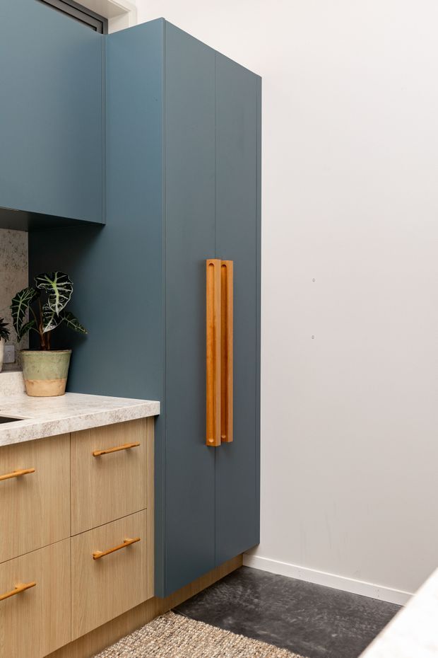 Close-up view of a contemporary kitchen featuring tall matte blue-grey cabinetry with long timber handles, light timber base drawers, a pale stone benchtop, and a dark textured floor. A potted plant sits in the corner, adding a natural accent to the restrained material palette.