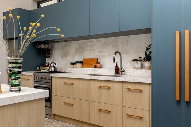 A contemporary kitchen with deep blue flat-panel overhead cabinetry and tall pantry doors, pale timber base cabinetry with long brass handles, and a light stone benchtop and splashback. A freestanding oven, stainless steel kettle, undermount sink with a curved mixer tap, open shelving, and decorative greenery complete the warm, refined scheme.