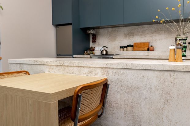 A contemporary kitchen with dark blue-green flat-panel cabinetry, a pale textured stone-look benchtop and backsplash, and a compact timber dining table with woven timber chairs in the foreground. A raised peninsula or island separates the dining area from the kitchen workspace, which includes a kettle, cooktop and small appliances. The palette is warm and neutral with contrasting dark cabinetry and natural timber details.