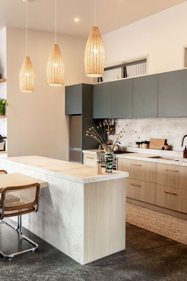 Contemporary kitchen with a large waterfall-edge island, integrated dark grey cabinetry, light timber lower cabinets, and a pale textured stone-look benchtop and splashback. Three woven pendant lights hang above the island, while bar stools, small appliances, and decorative branches add warmth. The space has dark polished concrete flooring and a warm neutral palette.