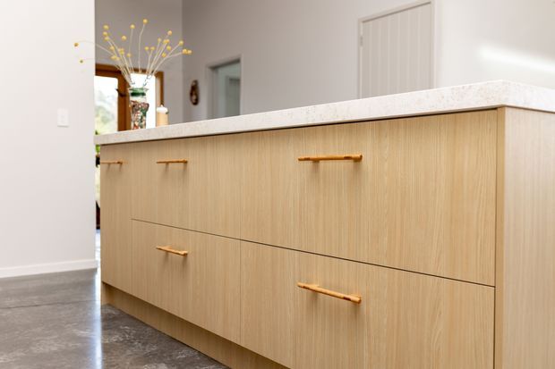 Close-up view of a contemporary kitchen island with light timber-grain cabinetry, slim brass or gold-toned pulls, and a pale stone benchtop. A grey polished concrete floor and light neutral walls create a clean, understated interior; a small glimpse of the adjacent living or circulation area is visible behind the island.