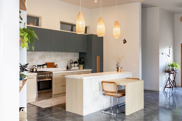 Bright contemporary kitchen with a large pale stone island and integrated dining extension, dark charcoal overhead cabinetry, light timber lower cabinetry, stainless-steel appliances, and three woven pendant lights. Polished concrete flooring and a high open connection to an adjacent living area are visible.