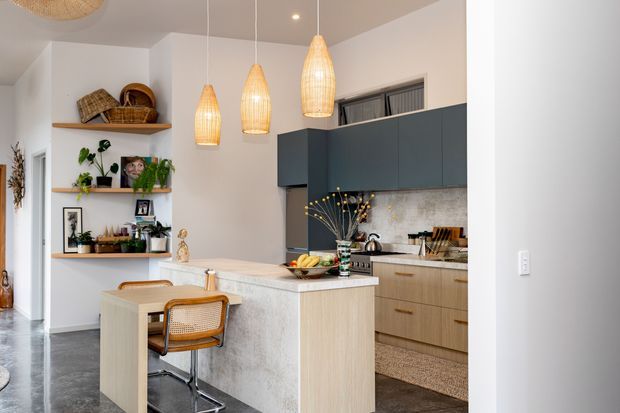 A bright contemporary kitchen with a compact island and integrated dining/work surface. The room features dark blue-grey upper cabinetry, light timber lower cabinetry, a pale textured stone-look island, open timber shelving with decor and plants, and three woven pendant lights. Dark flooring and warm wood accents add contrast to the light neutral walls.