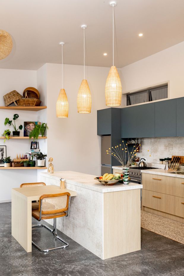 A bright, contemporary kitchen and dining area with a central island, pale timber cabinetry, dark blue-grey overhead cabinets, and a light stone benchtop. Three woven pendant lights hang over the island, while open timber shelves display plants, baskets, framed photographs, and decorative objects. A small dining table with cane-and-chrome chairs sits beside the island. The room has white walls, a warm neutral ceiling, a textured grey floor, and a softly mottled splashback.