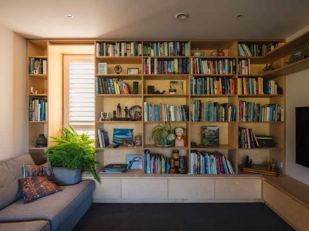 A warm, built-in floor-to-ceiling timber library wall occupies the room, with extensive open bookshelves, display niches, lower cabinetry and a recessed television area. A grey upholstered sectional sofa, patterned cushions, potted greenery and a window with white horizontal blinds are visible in the foreground. The space has a low, smooth painted ceiling with recessed downlights and a dark carpeted floor.