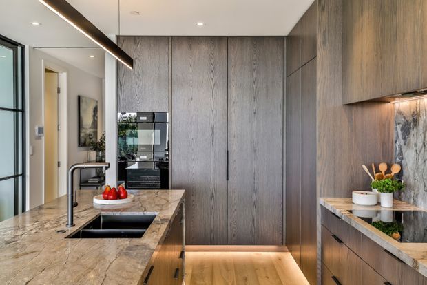 A refined contemporary kitchen featuring extensive dark timber-grain cabinetry, a large marble-look island with integrated black sink and tap, built-in black appliances, warm under-cabinet and toe-kick lighting, and a dramatic veined stone splashback. The space has a clean, sophisticated and warmly lit atmosphere.